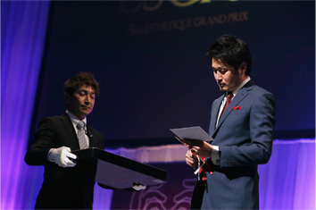 グランプリ受賞者の名前が書かれた封筒を受け取り、ハサミで開封する志田理事長。